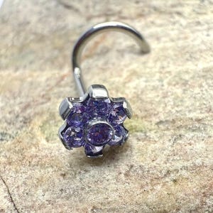 May include: A silver nose stud featuring a flower design. The flower is embellished with small, round, purple gemstones. The curved post is silver. The jewellery is set against a textured, light-coloured surface.