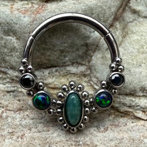 May include: A silver septum ring with an oval green gemstone at its centre, encircled by small silver beads. The ring also features black and iridescent green gemstones.