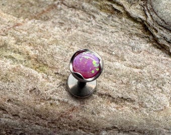 Puede incluir: Un pequeño pendiente labret plateado con una piedra de ópalo rosa redonda. El ópalo tiene motas verdes y está engastado en un bisel festoneado. El pendiente se muestra sobre una superficie de piedra texturizada de color marrón claro.