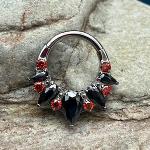 Peut inclure: Un anneau de septum argenté avec un design décoratif. L'anneau est orné de pierres précieuses noires et rouges. Les pierres précieuses sont disposées symétriquement, avec des pierres noires en forme de goutte et des pierres rondes rouges.