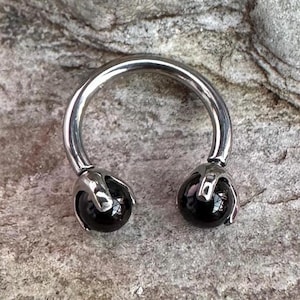 Pode incluir: Uma barra circular de prata com bolas de pedras preciosas pretas em cada extremidade. A barra é um tipo de joia corporal.