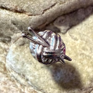 May include: A silver-colored snail-shaped charm with a ball of yarn on its back and knitting needles as antennae. The charm is highly detailed and sits on a textured, light-colored surface.