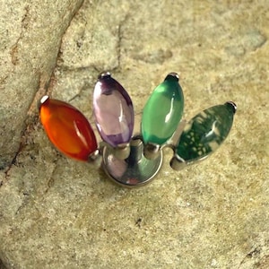 Könnte beinhalten: Ein dekoratives Schmuckstück mit vier farbigen, oval geformten Edelsteinen. Die Edelsteine sind orange, lila, grün und grün mit weißen Mustern, auf einer silbernen Basis. Das Stück wird auf einer strukturierten, hellbraunen Oberfläche präsentiert.