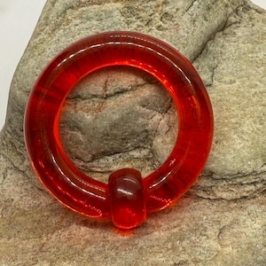 May include: A vibrant red, circular body jewelry piece. The piece is made of a translucent material and features a rounded, cylindrical bar. The jewelry is set against a textured, gray stone background.