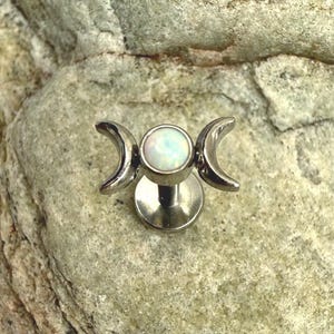 Könnte beinhalten: Ein silberfarbenes Lippenpiercing mit einem weißen Opal-Mittelstein und Halbmond-Akzenten. Der Schmuck ist vor einem strukturierten, grauen Steinhintergrund platziert.