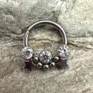 May include: A silver-colored hinged septum ring with three clear, round, sparkling gemstones and two silver star charms. The ring also features a cluster of small, silver-colored beads.