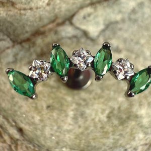 Op de afbeelding: Een zilveren oorbel met een gebogen ontwerp. Het heeft afwisselend groene marquise-geslepen edelstenen en vierkante heldere edelstenen. De oorbel is geplaatst tegen een getextureerde, neutrale achtergrond.