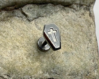 May include: A silver-colored coffin-shaped earring stud with a cross design. The earring is set against a textured, light-colored stone surface. The earring post is visible.