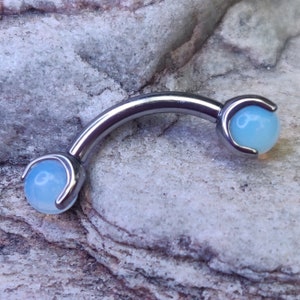 May include: A silver curved barbell with two light blue opal gems. The barbell is designed for body piercing.