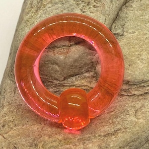 May include: A translucent orange captive bead ring, a type of body jewelry, with a circular design and a small bead. The ring is made of a clear material and is set against a textured stone background.