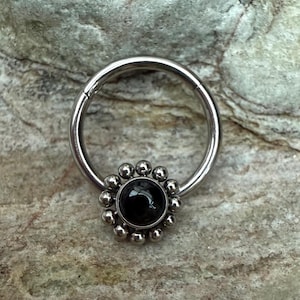 Könnte beinhalten: Ein silberfarbener Nasenring mit einem schwarzen Edelstein in der Mitte, umgeben von kleinen, silberfarbenen Perlen. Der kreisförmige Ring scheint aus Metall zu sein. Der Schmuck ist vor einem strukturierten, grauen Hintergrund platziert.