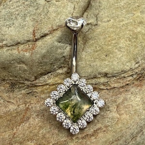 May include: A silver belly button ring featuring a clear, round gem and a square moss agate stone, encircled by small, clear gems. The moss agate displays green and yellow inclusions. The ring is designed for body piercing.