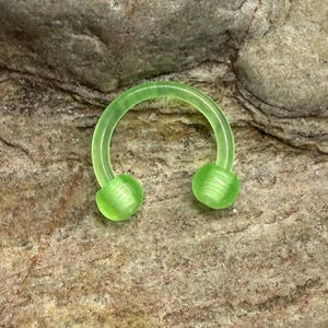 May include: A translucent green horseshoe-shaped body jewelry piece. The ends have rounded, ribbed balls. The jewelry is set against a textured, brown and gray stone background.