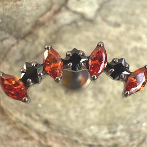 Könnte beinhalten: Ein silberfarbener Körperschmuck mit einer Reihe von orangefarbenen und schwarzen Edelsteinen im Marquiseschliff. Die Edelsteine sind in einem gebogenen Muster angeordnet, mit einer zentralen, runden, irisierenden Perle. Für Piercings.