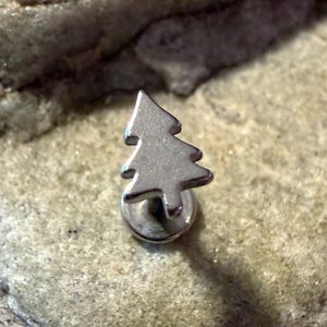 May include: A silver-coloured, tree-shaped stud earring. The earring has a flat back and a small, detailed tree design. The earring is set against a textured, light-coloured background. The earring is small and delicate, suitable for everyday wear.