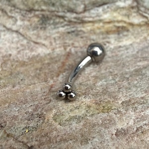 Può includere: Un piercing per sopracciglia curvo, color argento. Il gioiello presenta una grande sfera a un'estremità e tre sfere più piccole all'altra estremità. Il piercing è mostrato su uno sfondo neutro e strutturato.