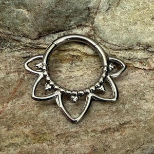 Könnte beinhalten: Ein silberner Septumring mit floralem Design. Der Ring hat einen Klappverschluss und ein dekoratives Element mit kleinen, runden Perlen und blütenblattförmigen Akzenten. Der Schmuck wird auf einer strukturierten, neutral getönten Oberfläche präsentiert.