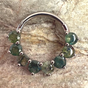 Könnte beinhalten: Ein silberfarbener Klappring, verziert mit einer Reihe grüner Edelsteine. Die Edelsteine sind rund geschliffen und mit kleinen silbernen Zinken gefasst. Der Ring wird vor einem strukturierten, hellbraunen Steinhintergrund gezeigt.