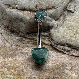 May include: A silver body piercing with a green gemstone heart-shaped charm. The charm is faceted and has a light green colour.