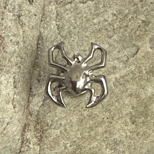 May include: A silver-colored spider-shaped pendant. The spider has a rounded body and eight legs, with a detailed design. The pendant is set against a textured, light gray background.
