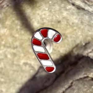 Puede incluir: Un dije en forma de bastón de caramelo con un contorno plateado. El bastón de caramelo presenta rayas de esmalte rojo y blanco alternadas. El dije es un accesorio festivo, perfecto para joyería o manualidades temáticas navideñas.
