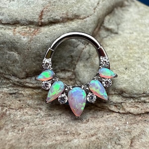 May include: A silver-colored septum ring featuring a row of teardrop-shaped opals in shades of pink, green, and white. The opals are accented with small, clear gemstones. The ring is designed for body piercing.
