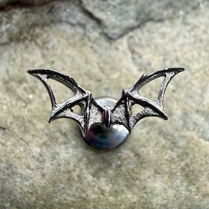 May include: A silver bat-shaped pin. The pin depicts a bat with detailed, outstretched wings. The wings have intricate detailing, and the pin is mounted on a circular base. The pin is likely made of metal.