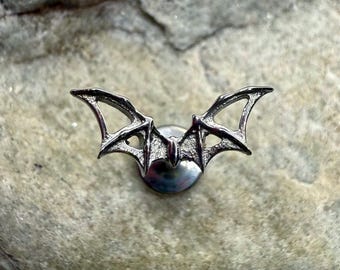 May include: A silver-colored bat-shaped pin. The pin features a detailed design of a bat with outstretched wings. The wings have intricate detailing, and the pin is mounted on a circular base. The pin is likely made of metal.