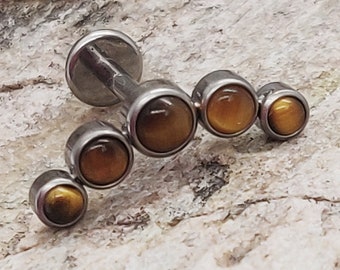 Puede incluir: Un piercing labial plateado con cinco piedras preciosas redondas de color ámbar. El piercing tiene una parte posterior plana y un poste con un diseño curvo. Las piedras están engastadas en biseles plateados.