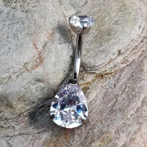 Puede incluir: Un piercing para el ombligo de barra curva en tono plateado con una pequeña piedra preciosa redonda y transparente en la parte superior y una piedra transparente más grande en forma de pera en la parte inferior. La barra une ambas piedras.