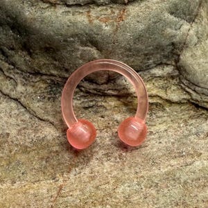 May include: A translucent pink horseshoe-shaped piercing jewelry with two rounded end balls. The jewelry is made of a smooth material and is set against a textured, gray and brown stone background.