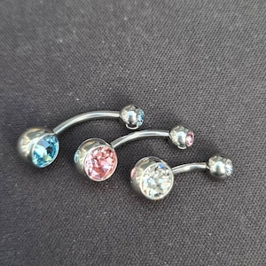 Könnte beinhalten: Drei silberne Bauchnabelringe mit verschiedenen farbigen Edelsteinen. Die Edelsteine sind hellblau, rosa und klar.