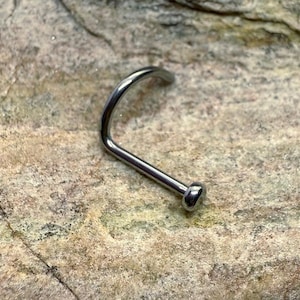 Può includere: Un piercing al naso color argento con un perno curvo e una piccola estremità arrotondata. Il gioiello è posto su una superficie di pietra strutturata, marrone chiaro. Questo pezzo è progettato per piercing al naso.