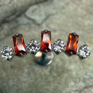 Può includere: Un orecchino argentato con una fila di pietre preziose rettangolari arancioni e rotonde trasparenti alternate. Le pietre preziose sono incastonate in un design lineare, creando un effetto scintillante. L'orecchino ha una chiusura a perno.