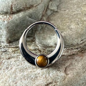 May include: A circular silver-toned metal segment ring featuring a polished finish and a single, round, golden-brown tiger's eye gemstone set at the bottom centre. The jewellery piece has a smooth, curved design with a hinged mechanism for closure.