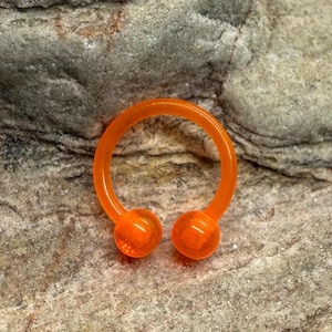 May include: An orange horseshoe-shaped body jewelry piece with two rounded ends. The jewelry is made of a translucent material and is set against a textured, gray stone background. This item is likely a septum or other piercing jewelry.
