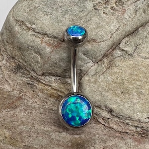 May include: A belly button ring with a silver-coloured metal stem and two round ends. Each end features a blue opal stone with a mix of blue and green hues. The jewellery is displayed against a textured grey background.