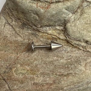 Peut inclure: Un piercing d'oreille en argent pointu, avec un dos plat et une pointe conique. Le bijou est présenté sur une surface de pierre texturée, brun clair. Conçu pour le perçage corporel.