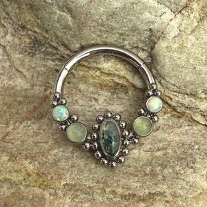 Könnte beinhalten: Ein silberner Septumring mit einem ovalen grünen Stein in der Mitte, umgeben von kleinen silbernen Perlen. Der Ring hat drei Paare kleinerer, runder, hellgrüner und schillernder Steine. Der Schmuck ist auf einem strukturierten, hellbraunen Stein platziert.