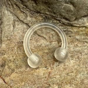 May include: A clear, horseshoe-shaped body piercing jewelry with two rounded ends. The jewelry is made of a translucent material and is set against a textured, light brown stone surface.