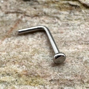 Puede incluir: Un aro de nariz de plata con una varilla curva en forma de L y un extremo redondeado.