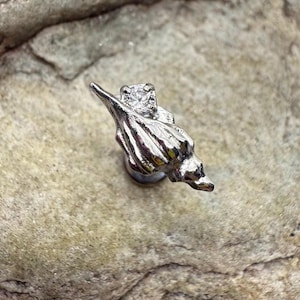 May include: A silver seashell stud earring with a small, square, clear gemstone. The earring is set against a textured, light-coloured stone surface. The earring's design is detailed and realistic.