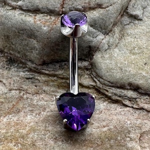 May include: A silver belly button ring with a purple heart-shaped gem on the bottom and a smaller purple round gem on the top.