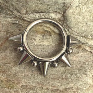 May include: A silver-colored, hinged septum ring with a series of conical spikes and small ball accents. The jewelry is designed with a punk rock aesthetic. The ring is displayed against a textured, neutral-toned background.