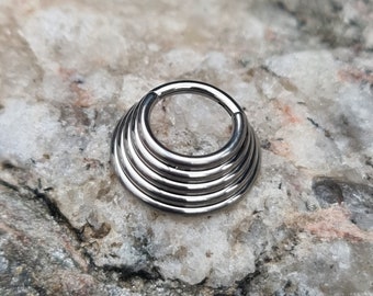 May include: A silver-colored, multi-layered nose ring. The jewelry features a circular design with multiple stacked rings, creating a layered effect. The nose ring is made of a shiny, metallic material and is set against a textured, light-colored background.
