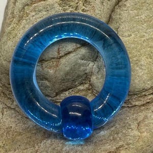 May include: A vibrant blue, circular body jewelry piece. The piece is made of a translucent material, with a small, rounded bead at the bottom. The jewelry is set against a textured, gray stone background.