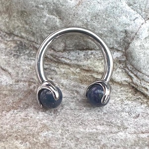 Pode incluir: Uma barra circular de prata com duas bolas de pedras preciosas azuis nas extremidades.