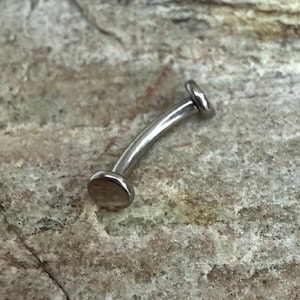 May include: A curved, silver-colored eyebrow piercing. The jewelry has a flat disc on each end of the curved bar. The piercing is shown against a neutral, textured background.