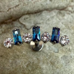 May include: A silver-toned ear piercing stud featuring a curved design. It has three rectangular, iridescent blue gems and four clear, round gems. The jewelry is set against a textured, light brown background.
