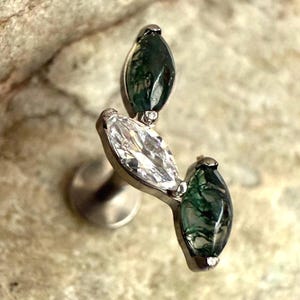 May include: A silver ear piercing stud featuring three marquise-cut gemstones. Two are dark green, and one is clear. The stud is set against a textured, light brown background.